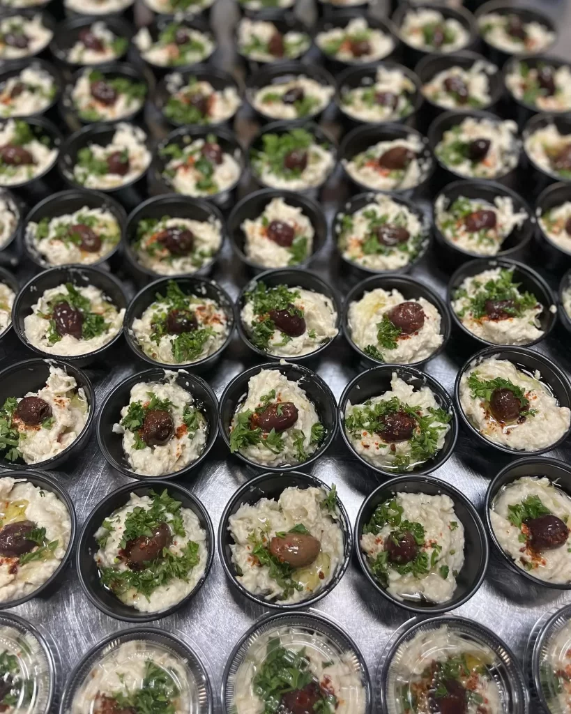 Rows of single-serve babaganoush cups garnished with olives and parsley for a catering order