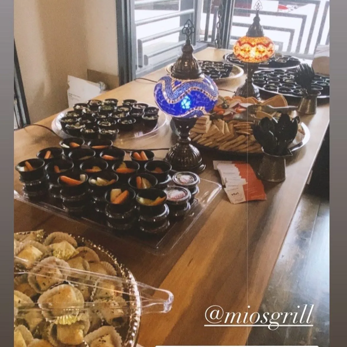 Catering spread on a wood table with trays of mezze dips, veggie cups, and spinach börek beside Turkish mosaic lamps